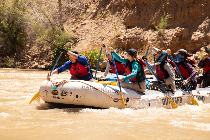 A group of people is enjoying a rafting adventure on a river. They are in an inflatable raft, paddling through the water with enthusiasm. The surrounding landscape features rocky cliffs and lush greenery, adding to the scenic beauty of the experience. The rafters are wearing life jackets, ensuring their safety during the activity.